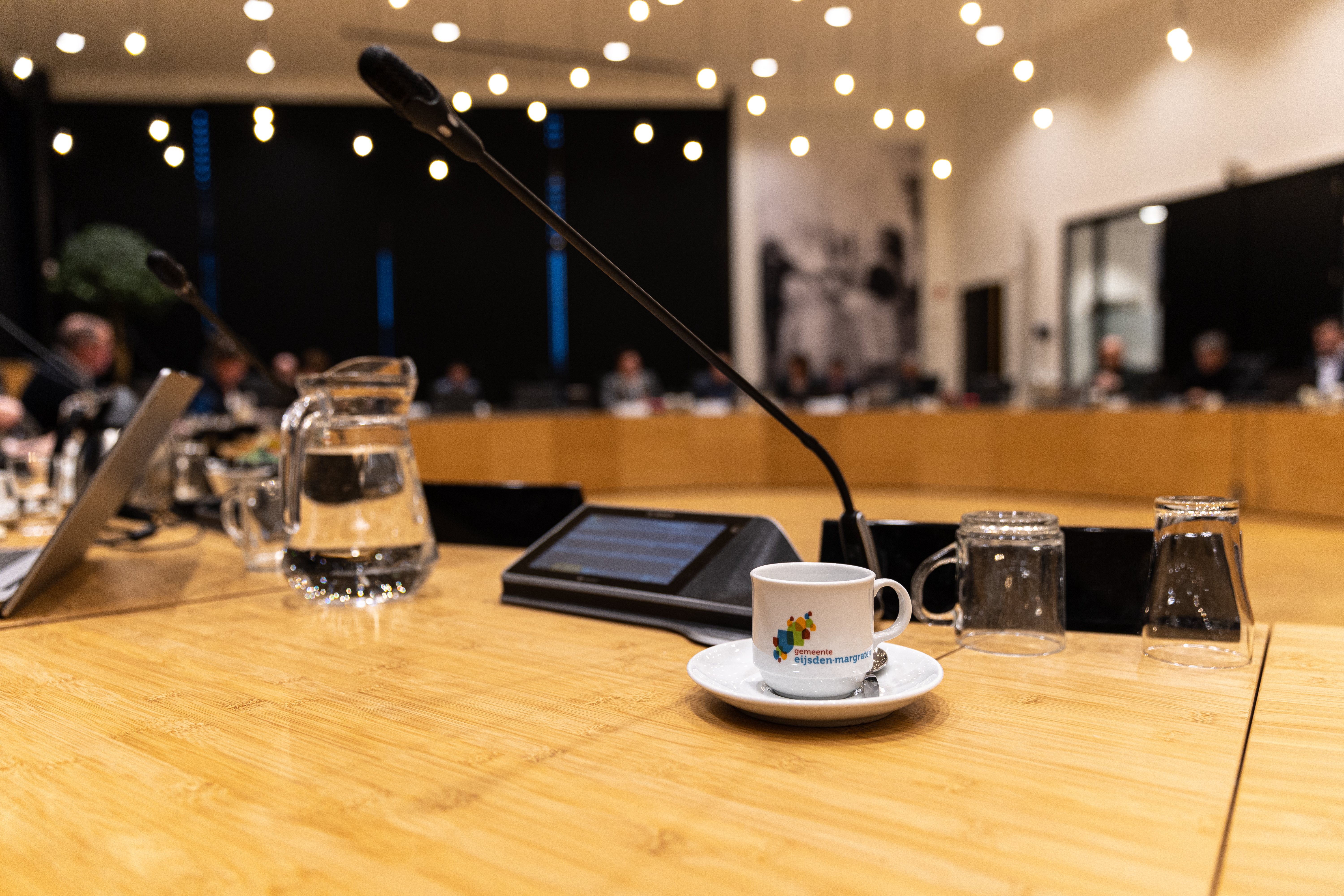 Tafel in raadzaal met koffiekopje, kan water, drinkglas, microfoon en scherm. 